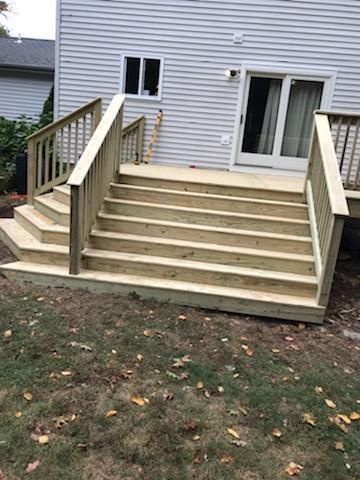 Wooden deck with stairs leading to a sliding door, flanked by railings. Steps are bare wood on a grassy lawn.