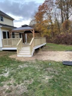 Wooden deck with gazebo, stairs, and railing, attached to a light yellow house, surrounded by grass and trees.