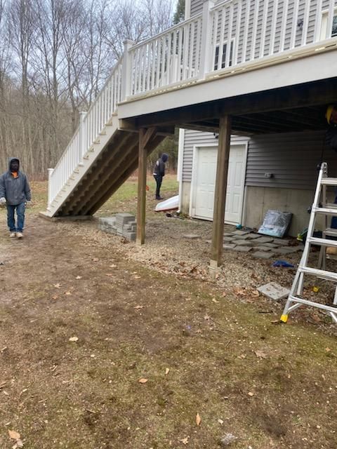 Deck with stairs, supported by wooden posts. Two people near the deck. Unfinished ground.