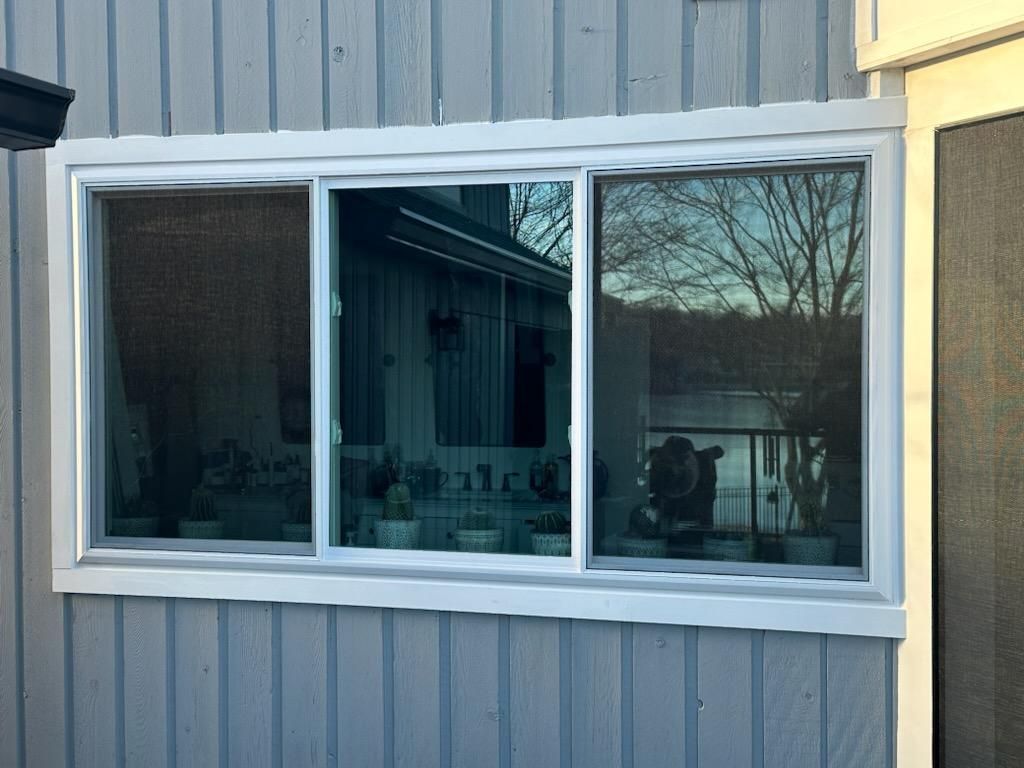 White-framed, three-pane window on a gray building exterior. The window reflects the outside surroundings, with a screen in the left panel.