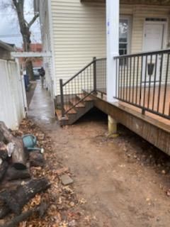 Side view of a house with a wooden deck and steps. A dirt path leads along the side of the house.