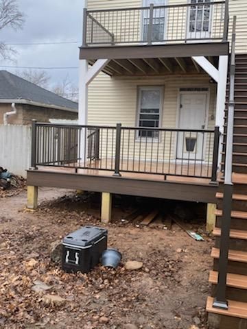 Two-story deck with brown composite deck boards, black railings, and a beige house. Ground has debris.