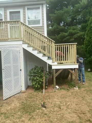 Wooden deck with stairs leading up to a house. A person stands on the deck, and another is nearby.