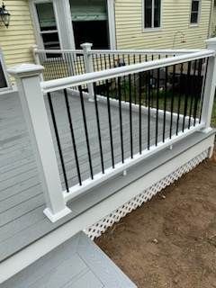 White deck railing with black vertical spindles. Gray deck boards. Yellow house in background.