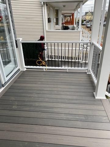 A porch with gray deck boards and white railing. A person in a red jacket holds a hose.