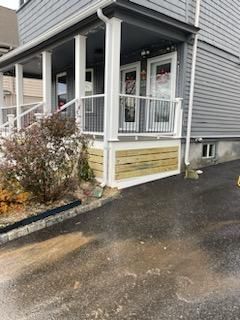 A house with a front porch. White railings and supports, light wood paneling, gray siding, and a dark asphalt driveway.