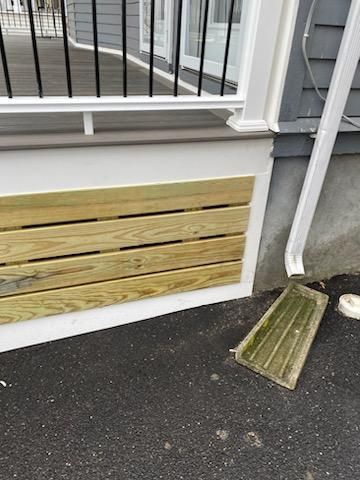 Wooden slats on the side of a deck, next to a white gutter and gray house.