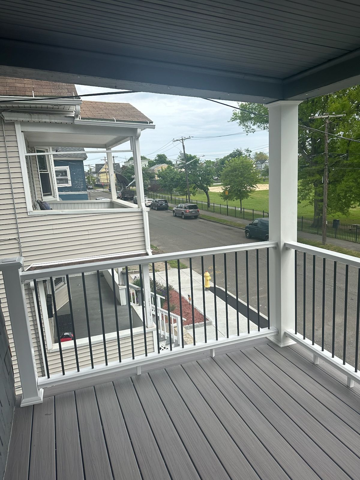 Balcony overlooking street with cars, trees, and blue sky. Black railing, grey deck, white supports.