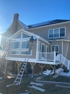 Construction worker on ladder, building a sunroom addition on a two-story house. White trim, beige siding, blue sky.