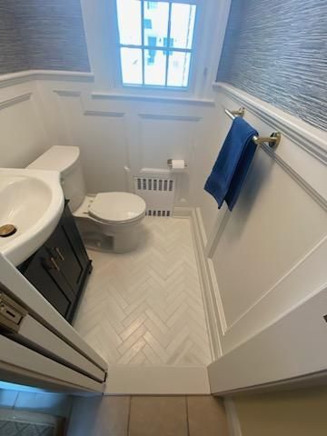 Small, white bathroom with herringbone floor. Toilet, sink, window, radiator, and towel rack.