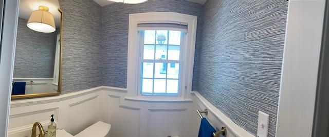 Bathroom with blue textured wallpaper, white wainscoting, gold fixtures, and a window.