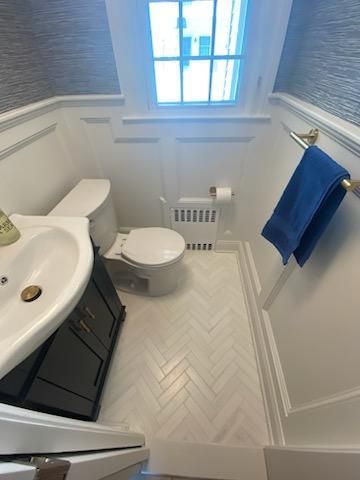 Small, white bathroom with a toilet, sink, and window. White herringbone tile floor and blue towel.