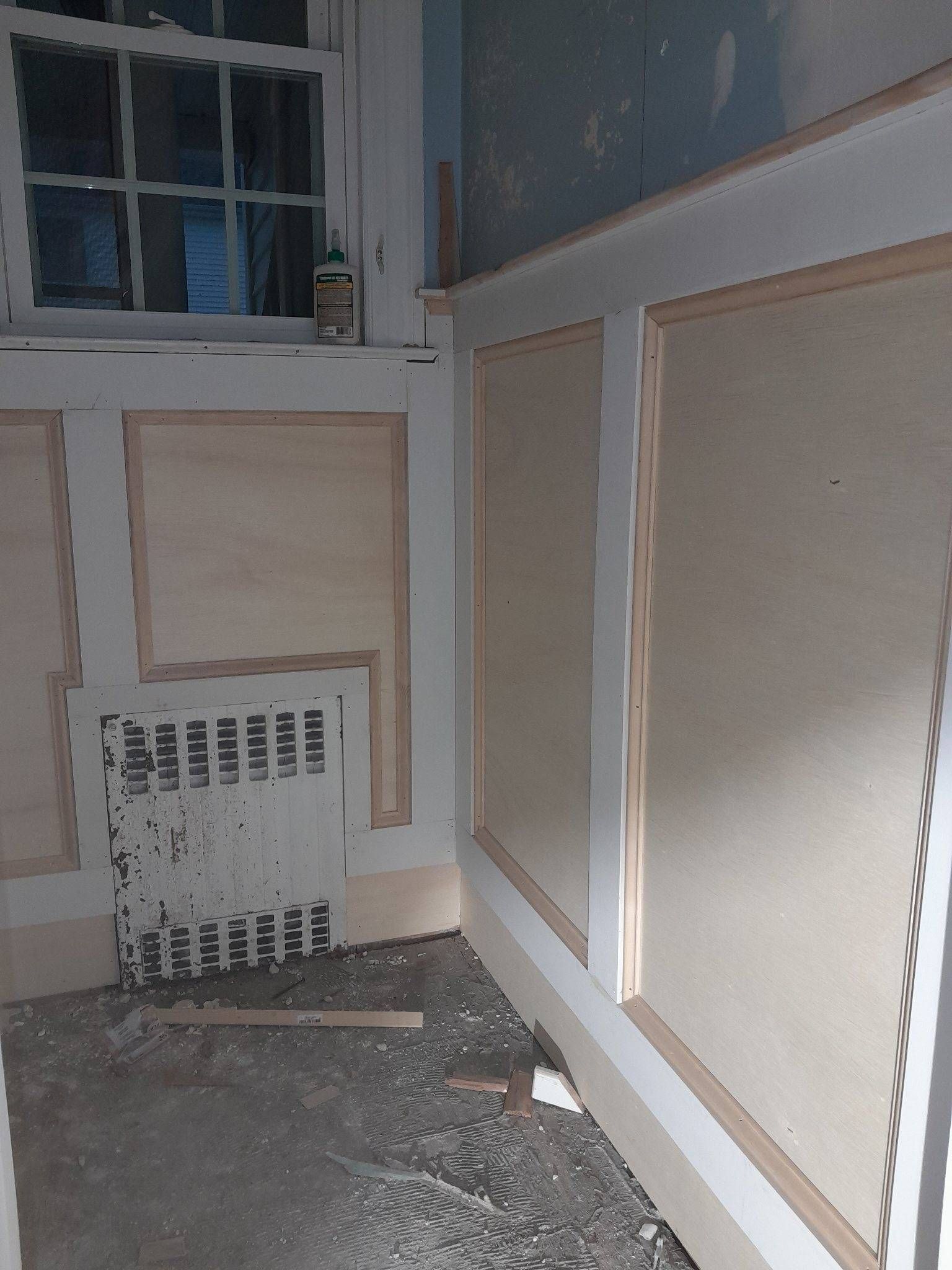 Room corner with wainscoting under construction; white trim, light panels, radiator, window.