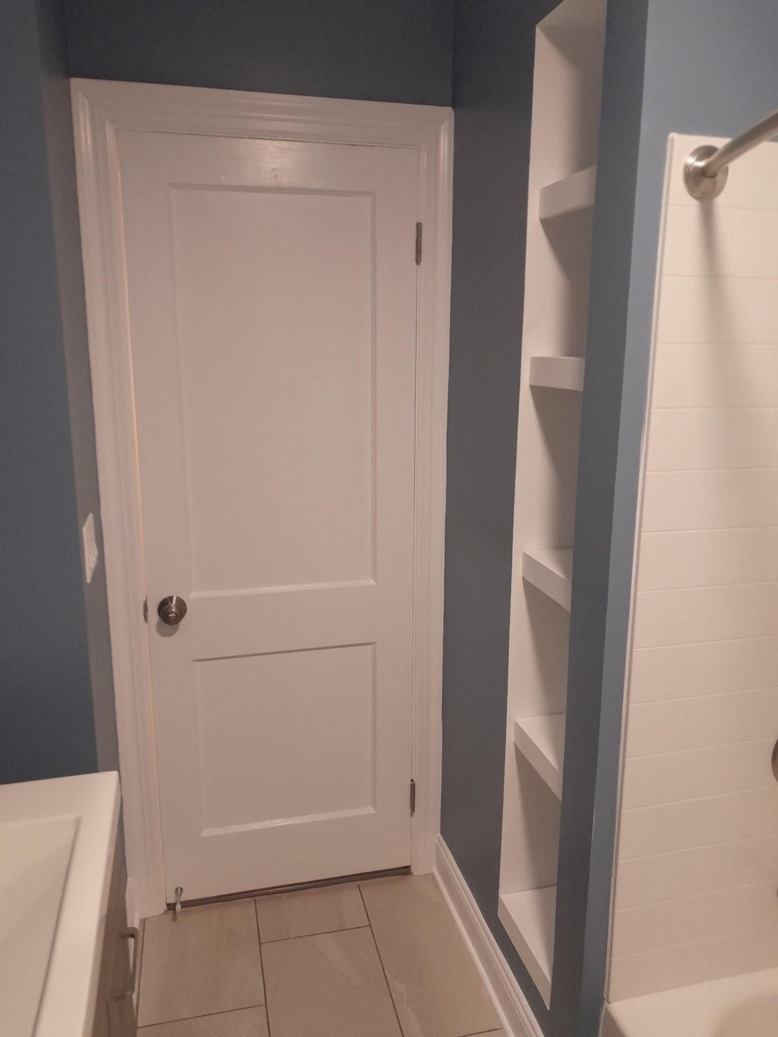 Bathroom with white door, built-in shelves, and shower. Blue walls and beige floor tiles.