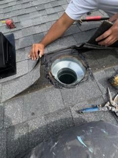 Person on a roof repairs a vent pipe flashing, lifting asphalt shingle. Black roofing material and tools visible.