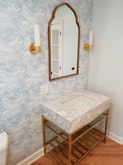 Bathroom with wave patterned wallpaper, a gold-framed mirror, marble sink on a gold stand, and sconces.