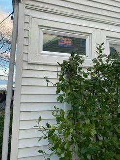 White house siding with a window, greenery in the foreground, and a sign visible in the window.
