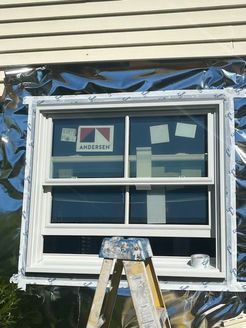 White window installed in a house, surrounded by silver flashing tape. A small ladder is in front.