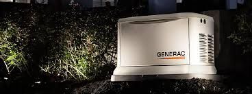 A Generac generator sits on a base surrounded by dark mulch and shrubs at night.