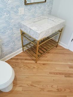 A marble-topped vanity with gold legs and a glass shelf in a bathroom with wave wallpaper.
