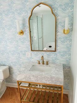 Bathroom with light blue wave wallpaper, gold-framed mirror, marble countertop, gold faucet, and gold sconces.