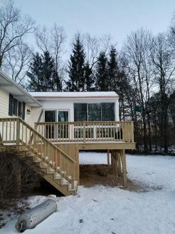 A deck with stairs attached to a house with a sunroom, with snow on the ground, and bare trees in the background.