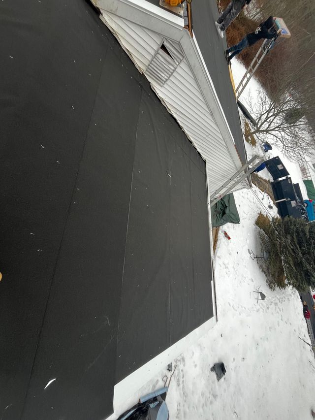 Black roofing on a white-sided house, surrounded by snow. A ladder and trees are visible in the background.