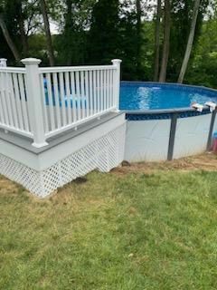 White deck and railing next to an above-ground pool filled with blue water; green grass in front.