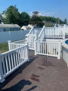 White railing surrounds a dark brown deck. A small section of above-ground pool is visible. In the background are houses and a white fence.