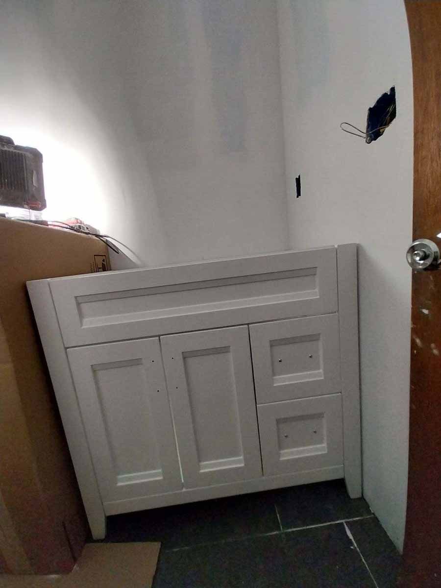 White bathroom vanity cabinet against a white wall; electrical box visible; brown door to the right.