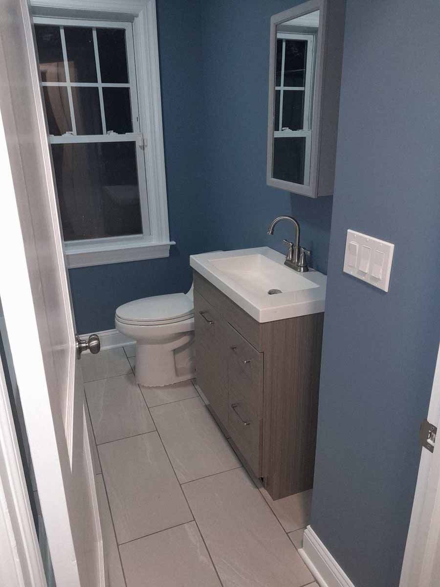 Small blue bathroom with a toilet, sink, mirror, and a window.