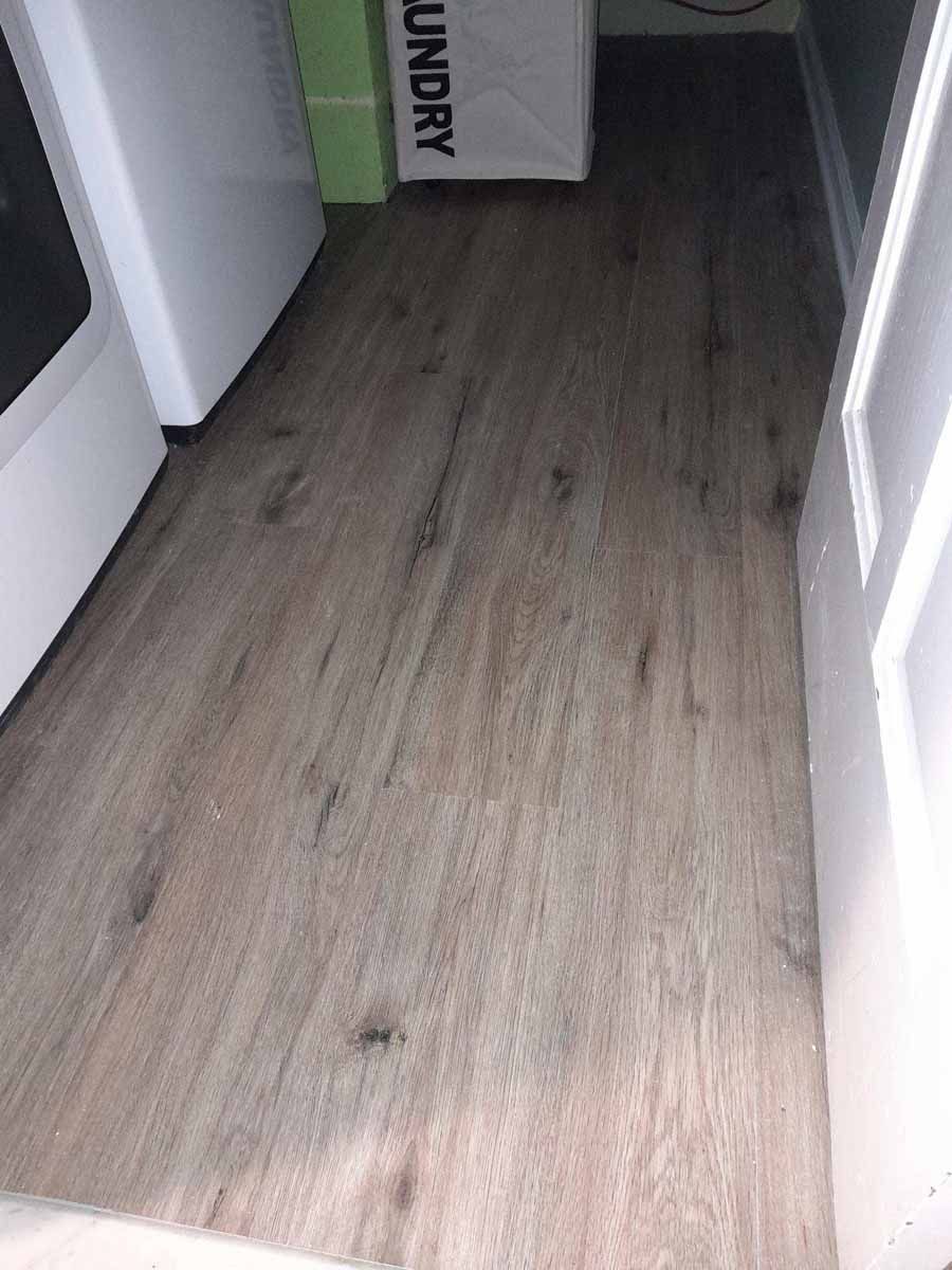 Wood-look flooring in a laundry room, between a white appliance and a white door. A laundry bag is in the background.