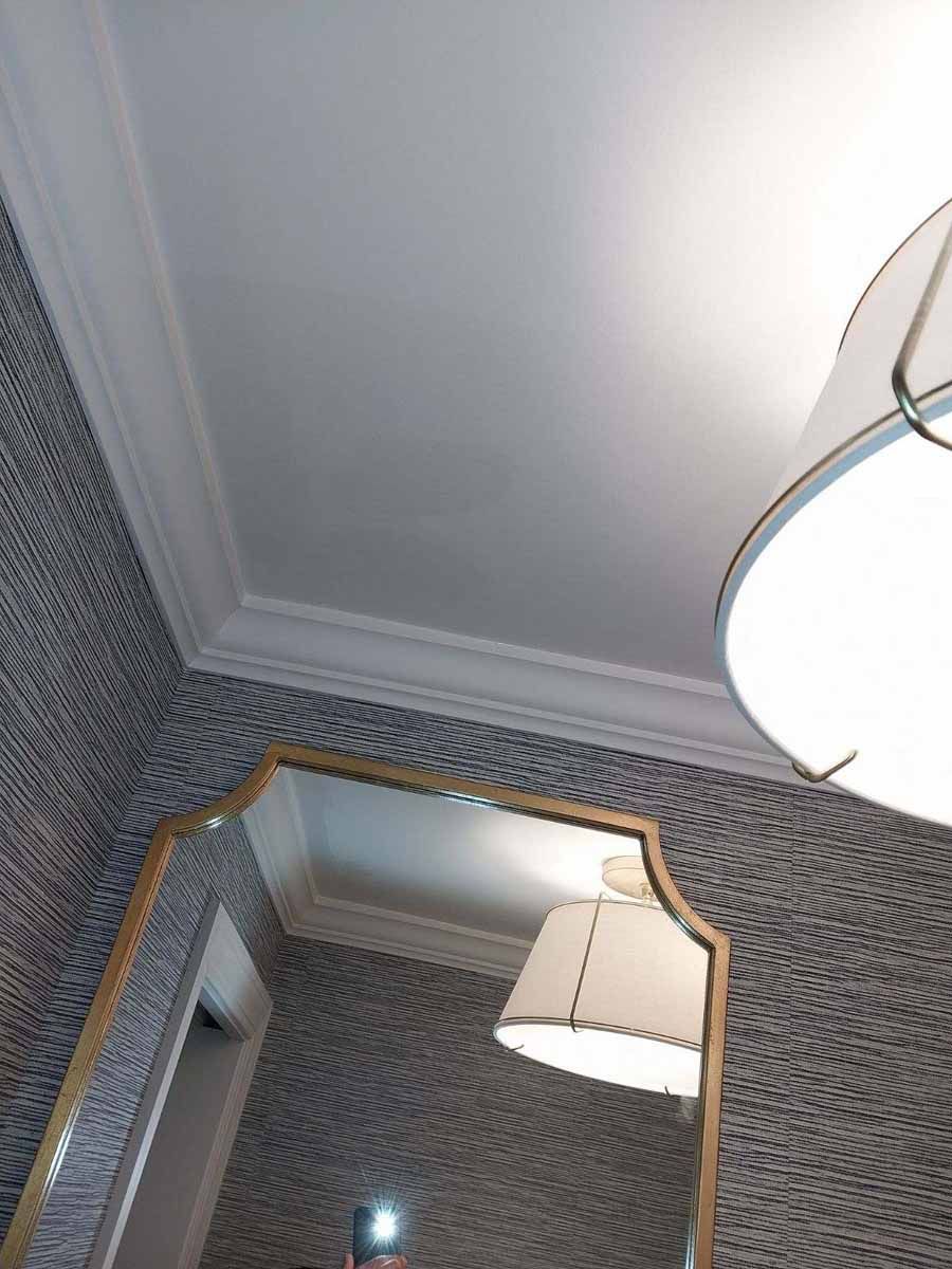 Gold-framed mirror reflects a lit lamp and textured wallpaper in a room with white crown molding and ceiling.