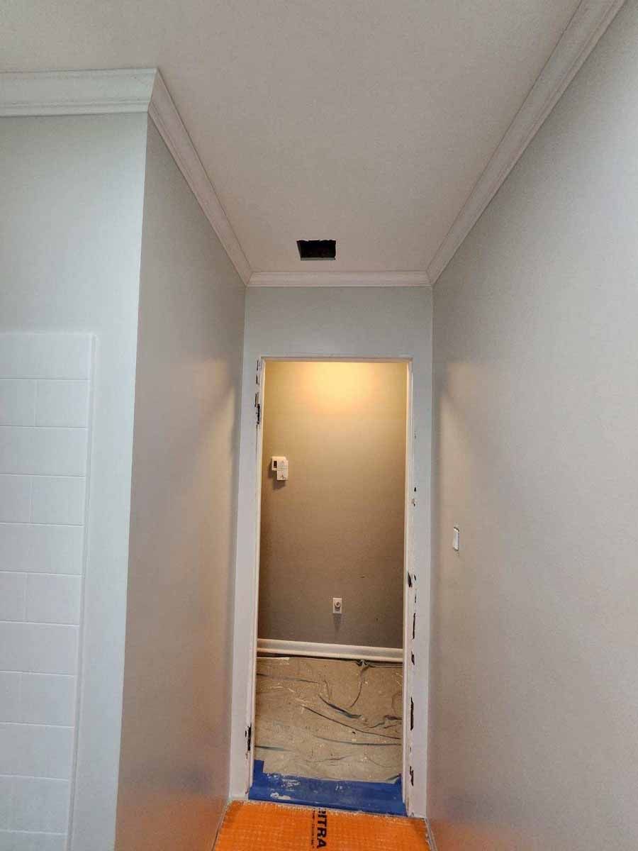Narrow hallway with white walls and trim, leading to a doorway. Orange floor in foreground, unfinished space within doorway.