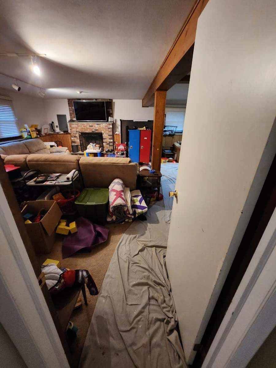 View of a cluttered room with furniture, toys, and a partially open door. Beige couch, fireplace and wood beam are visible.