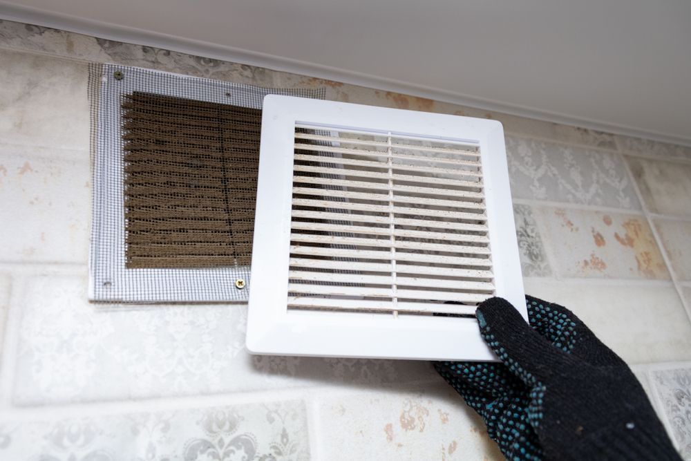 Person wearing glove removing a dirty bathroom vent cover from a wall.