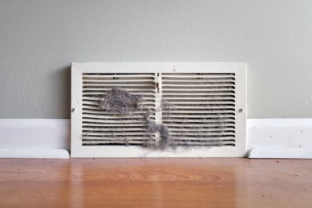 Dusty air vent on a gray wall above wooden flooring.