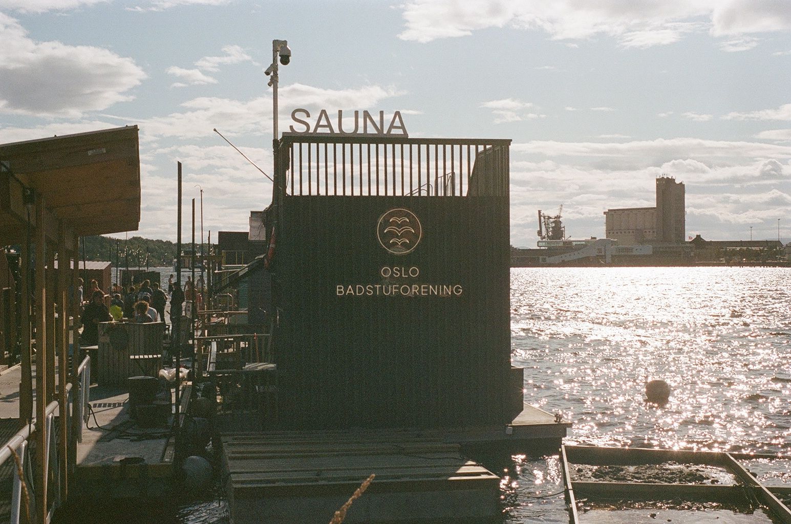 Wooden sauna hut on a dock by shimmering water at sunset, with a distant harbor skyline
