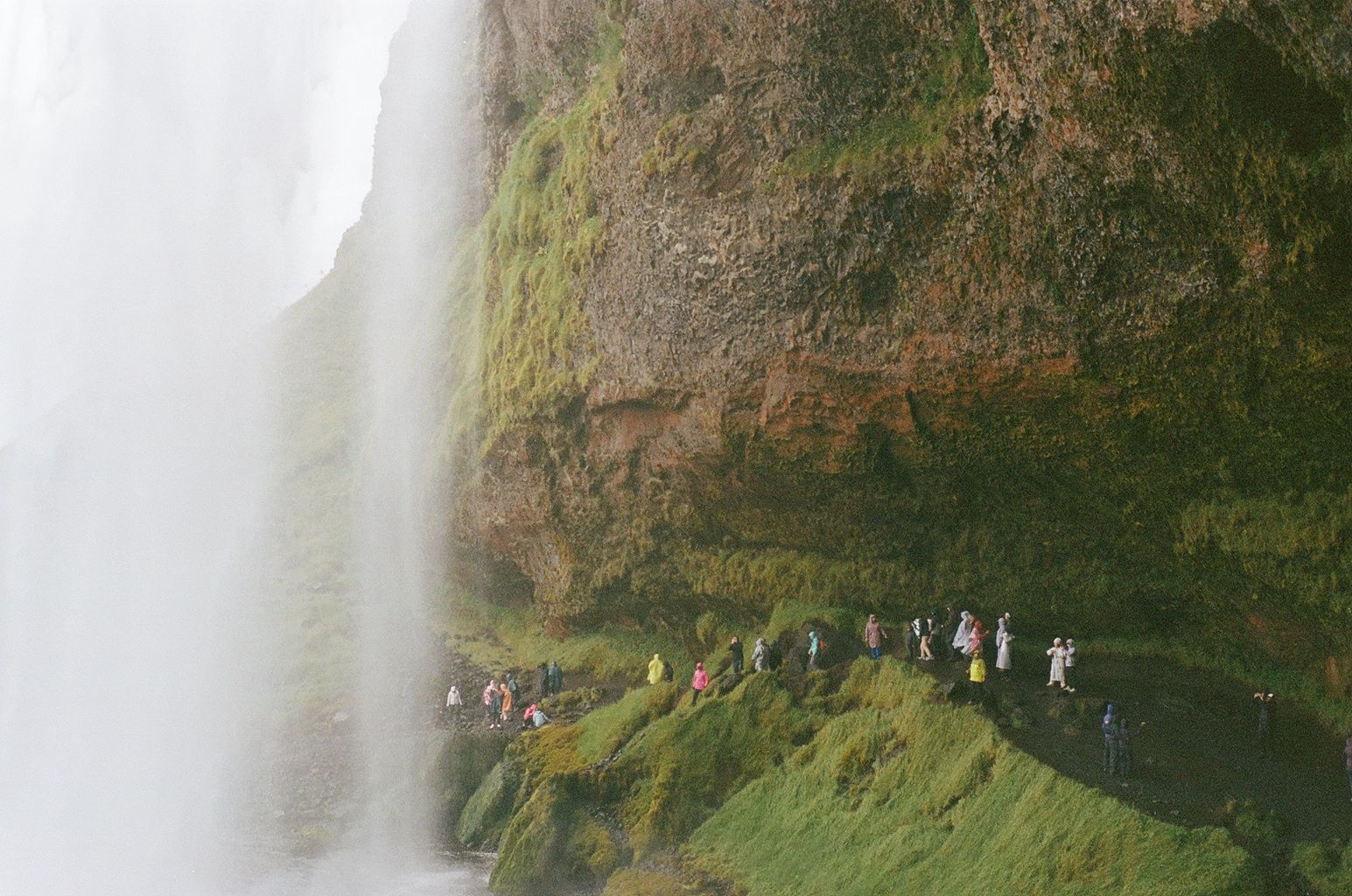 Waterfall beside a cliff with tourists walking along a narrow green path beneath the mist