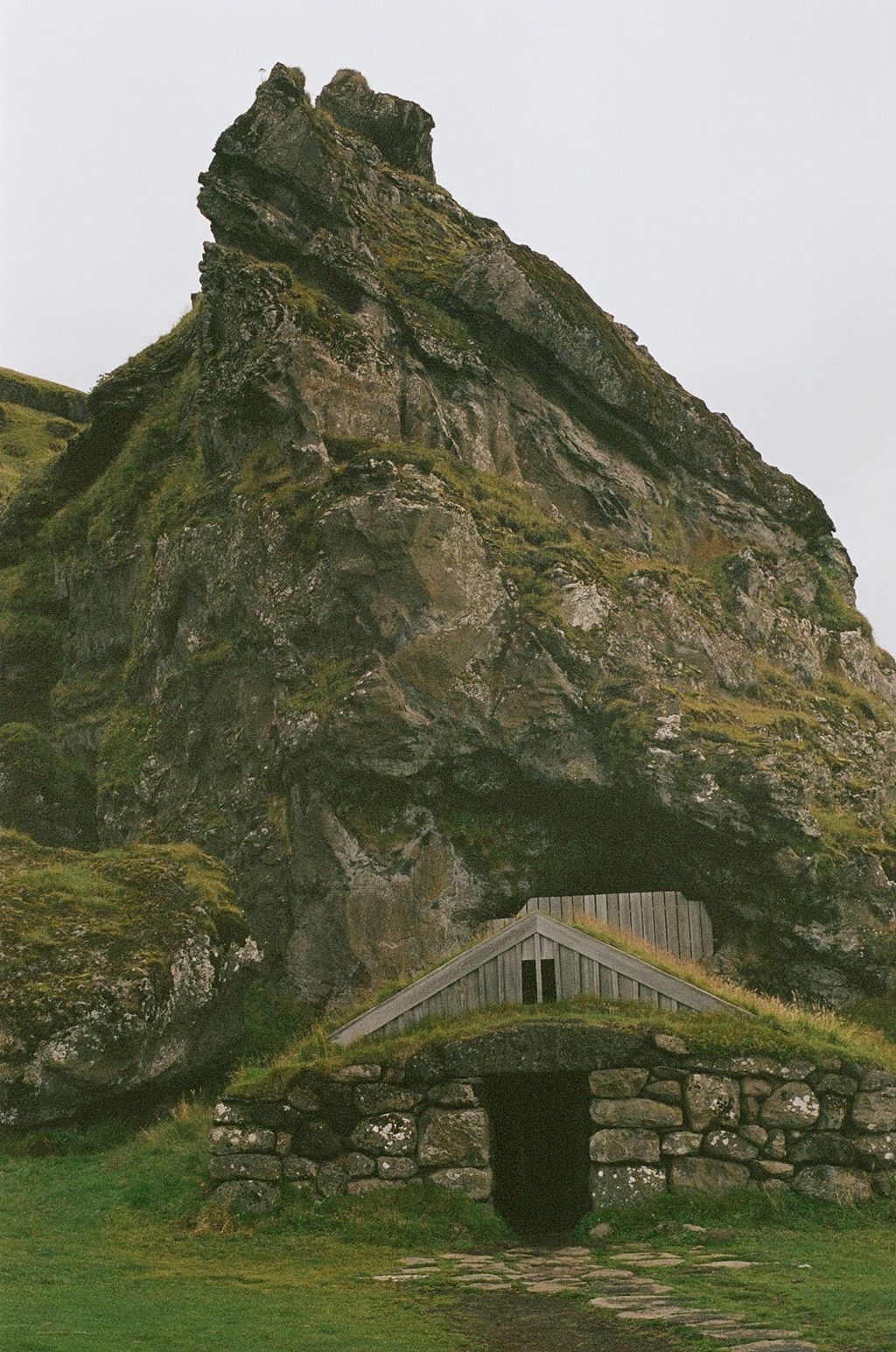 Rocky green cliff towering above a small stone hut with a grassy roof.