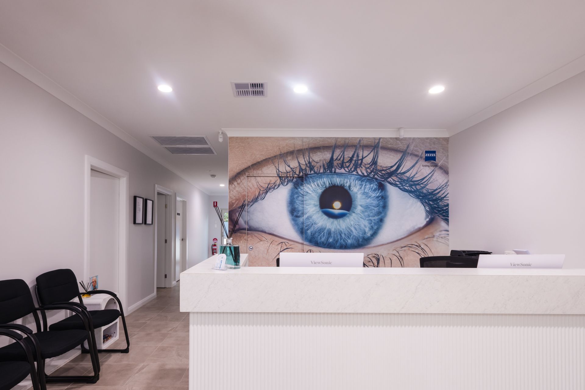 Bright clinic reception with white desk, black chairs, and a large blue eye mural on the back wall.