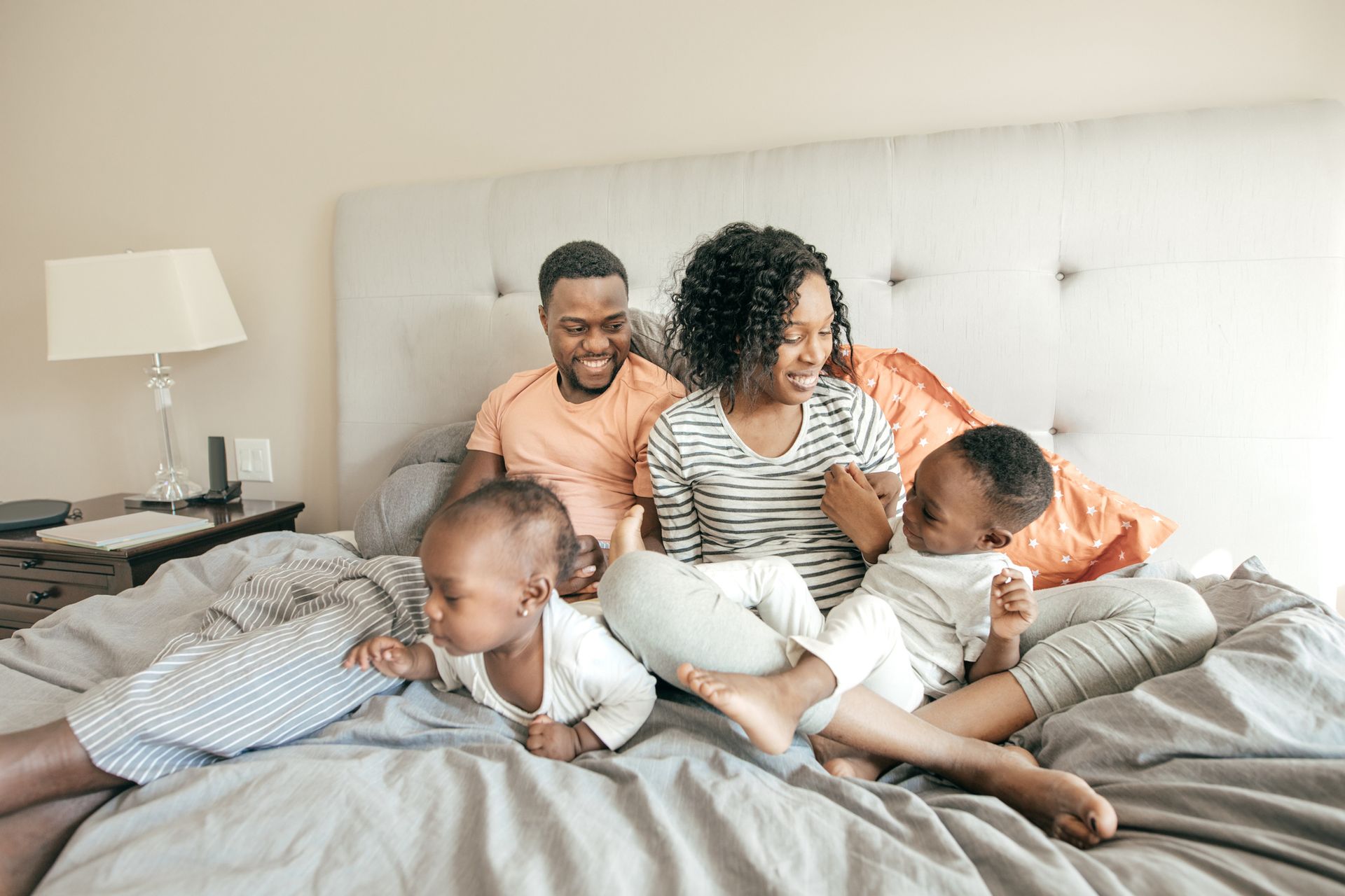 Family of four on a bed, smiling. Adults with two toddlers.