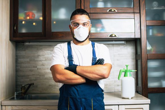 Person in protective gear with arms crossed, standing in a kitchen with a spray bottle.
