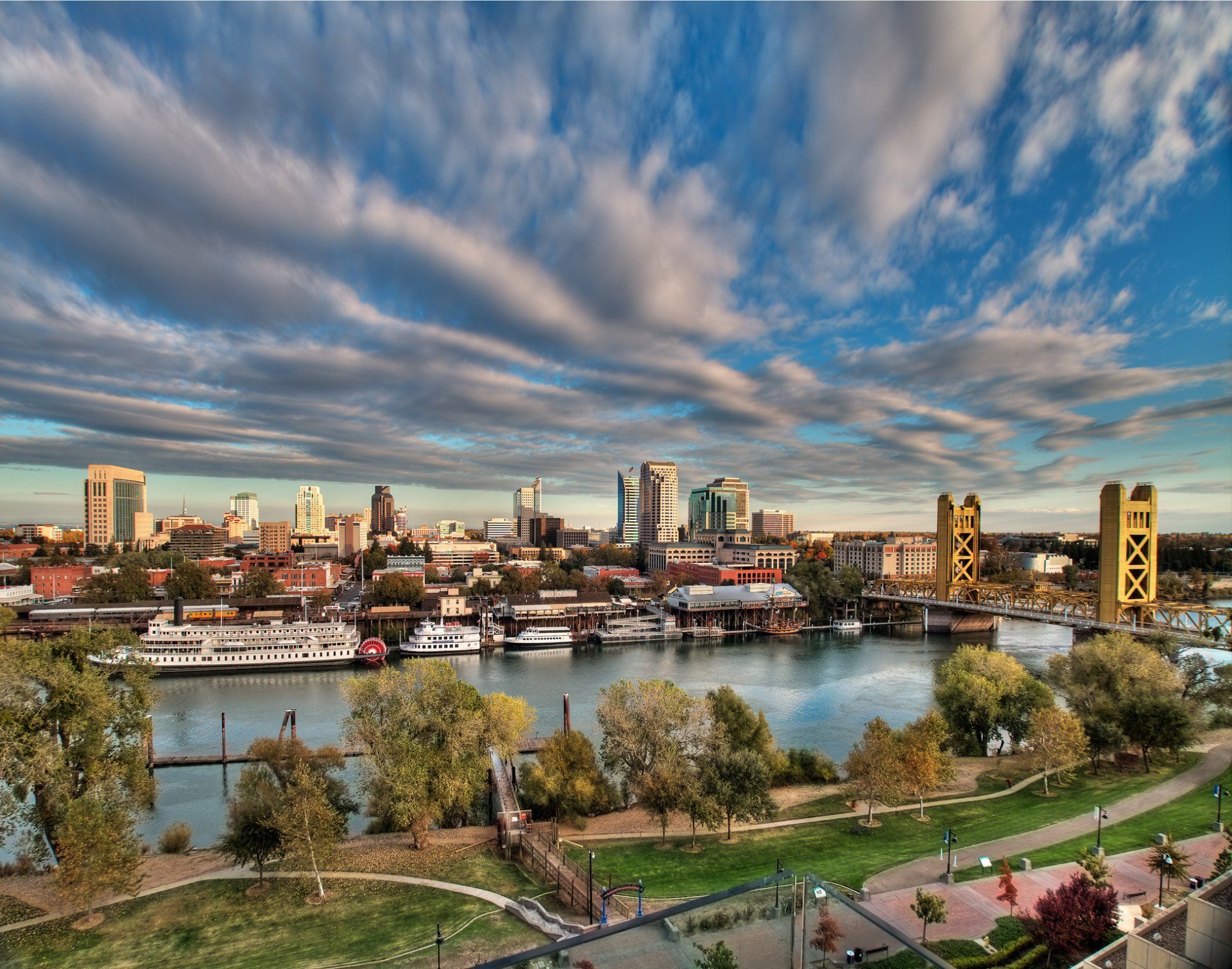Sacramento, CA skyline with the Tower Bridge in gold, river, boats, and cloudy blue sky.