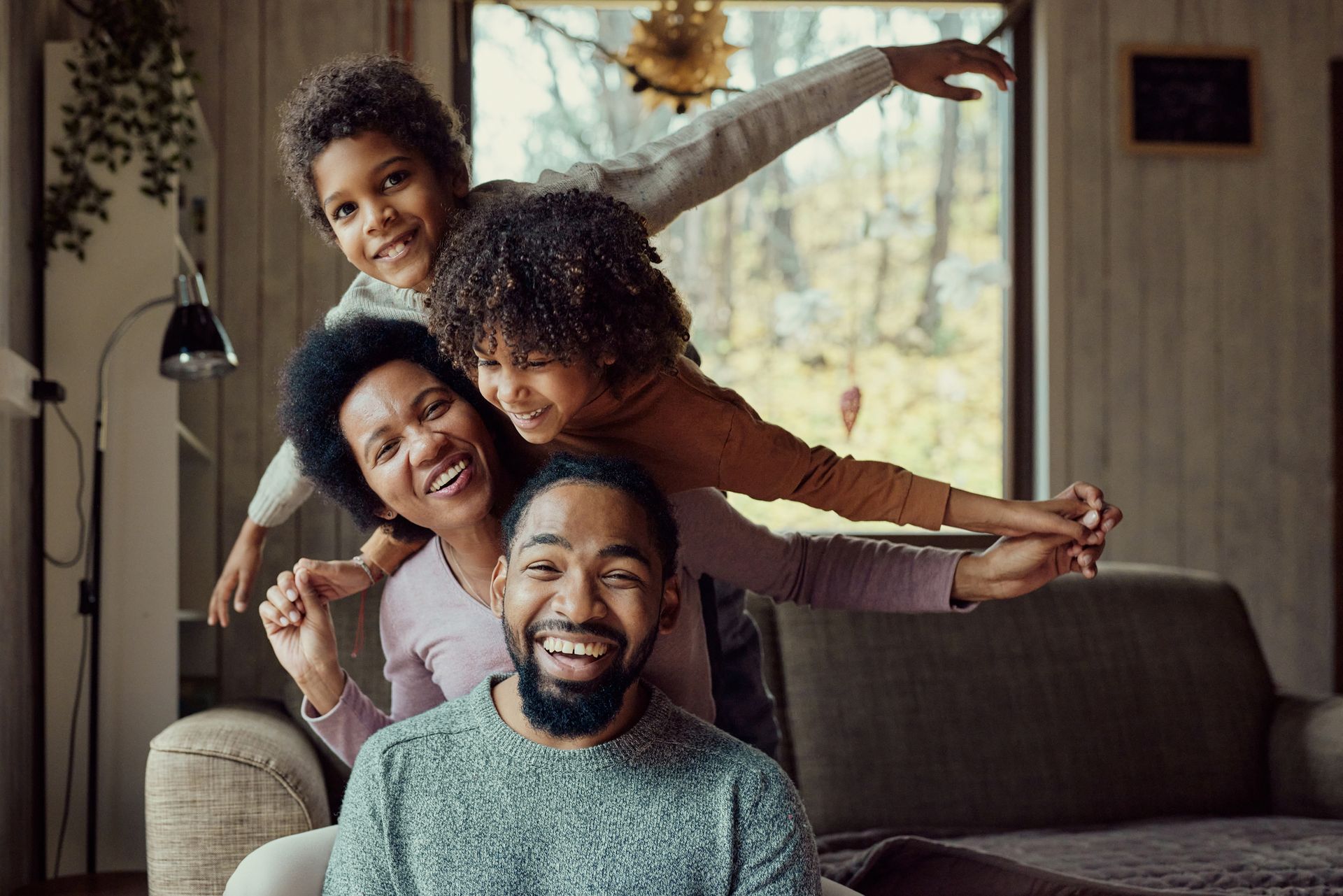 Family of four smiling, stacked together, indoors, arms outstretched, near a window.