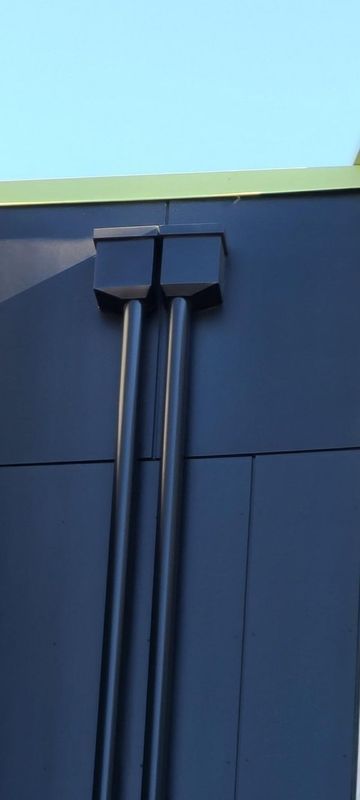 Two dark pipes descend from a box on a dark building wall, with a blue sky background.
