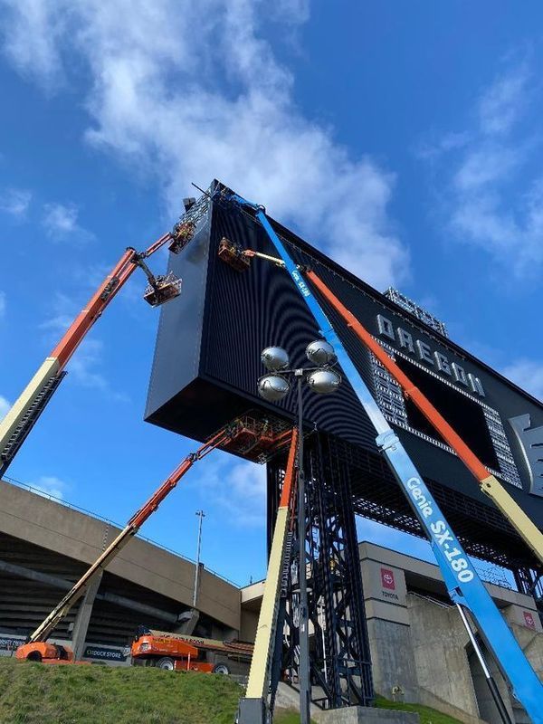 Several boom lifts working on a large black scoreboard against a blue sky. Stadium in background.