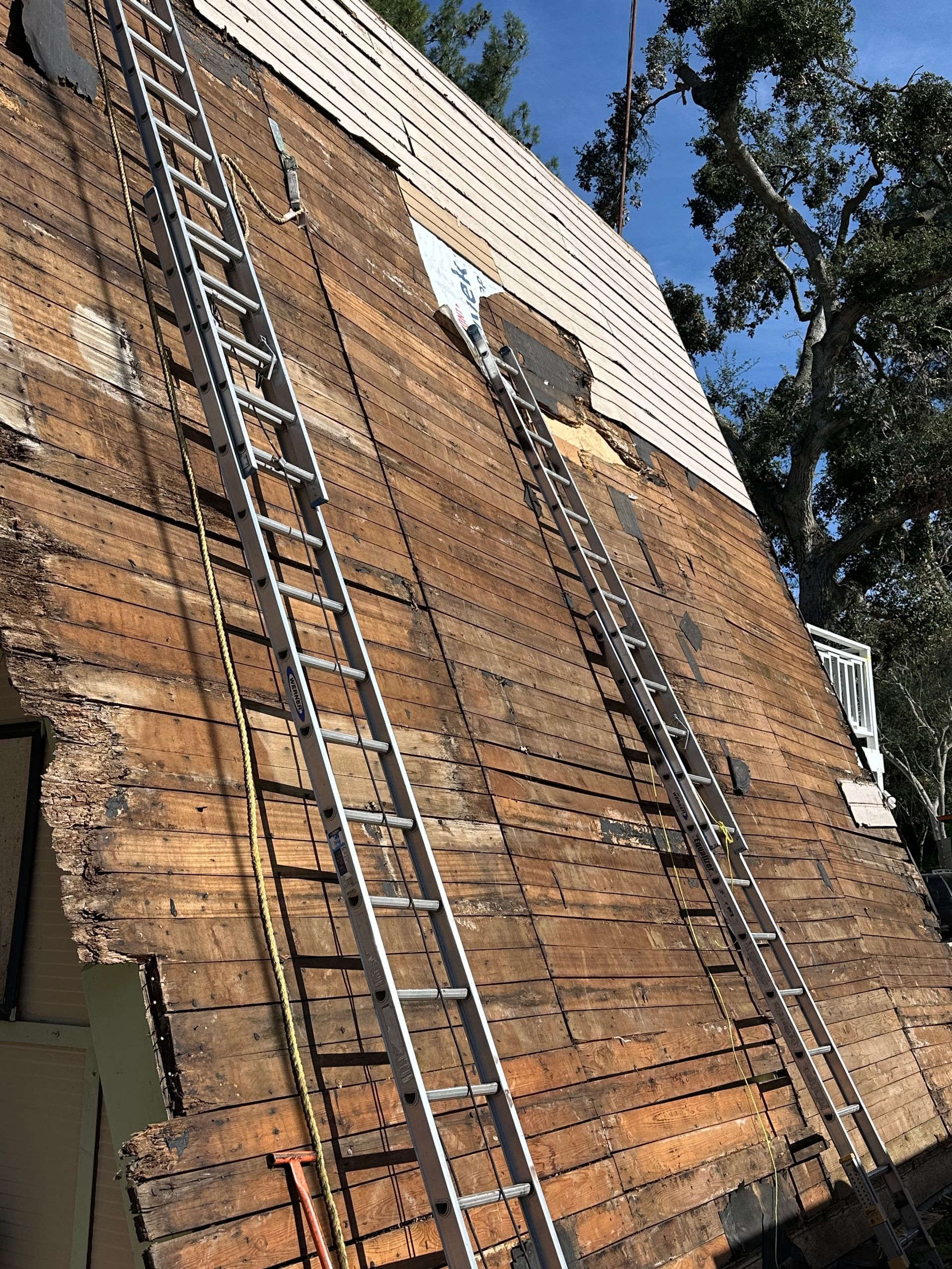 Two ladders on a roof with exposed wood, partially missing shingles, and a clear sky.