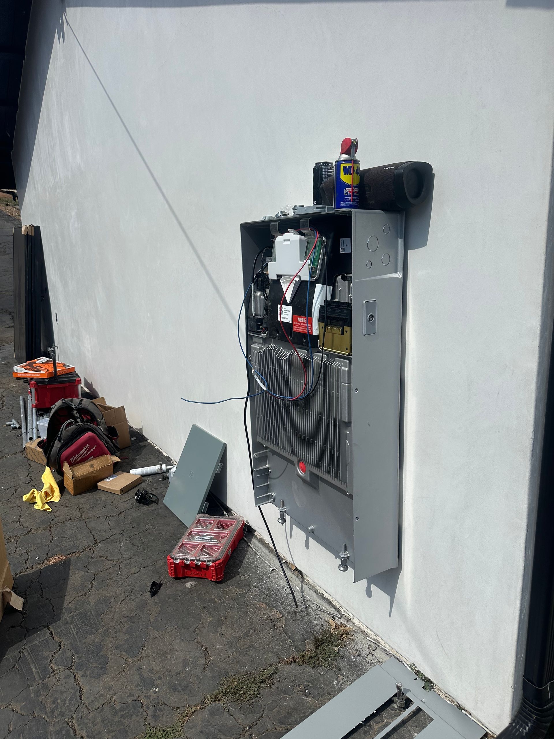 Open electrical panel mounted on a white exterior wall, with tools and debris on the ground nearby.