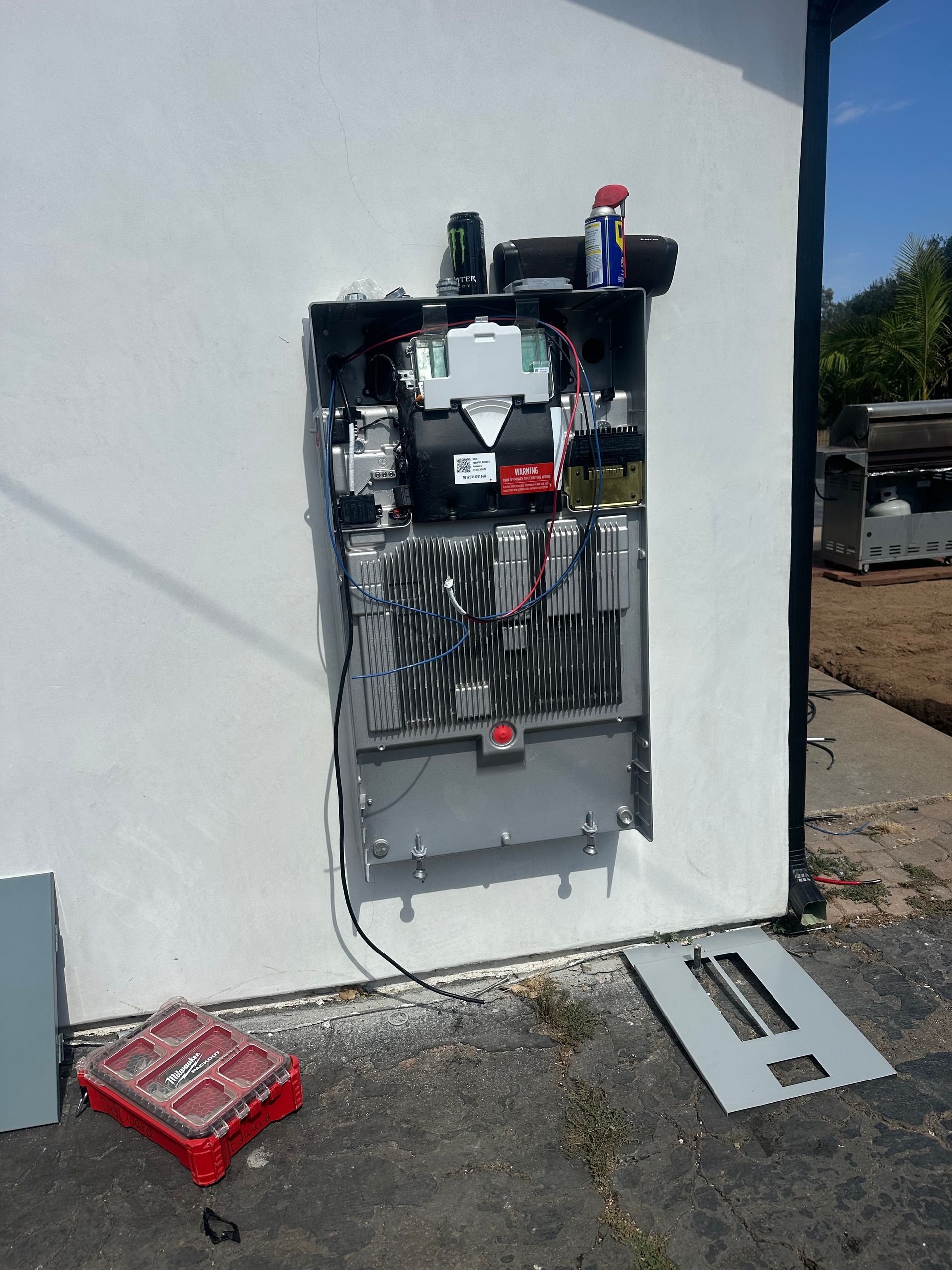 Open electrical panel mounted on a white exterior wall, with tools and red parts on the ground nearby.